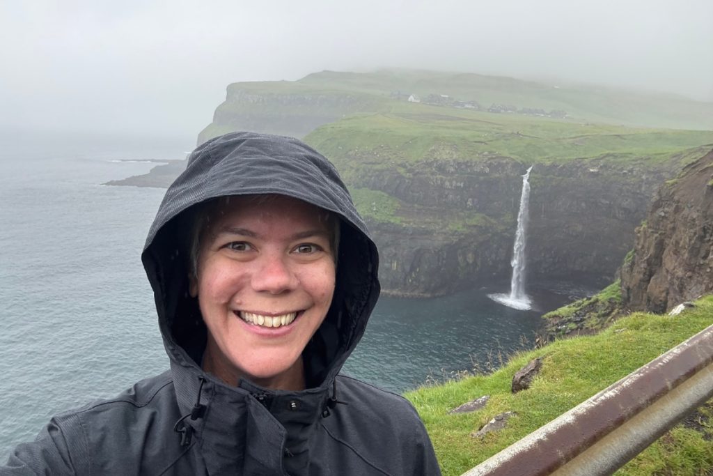 A woman in a rain jacket smiling in front of Múlafossur waterfall on a foggy day, Faroe Islands