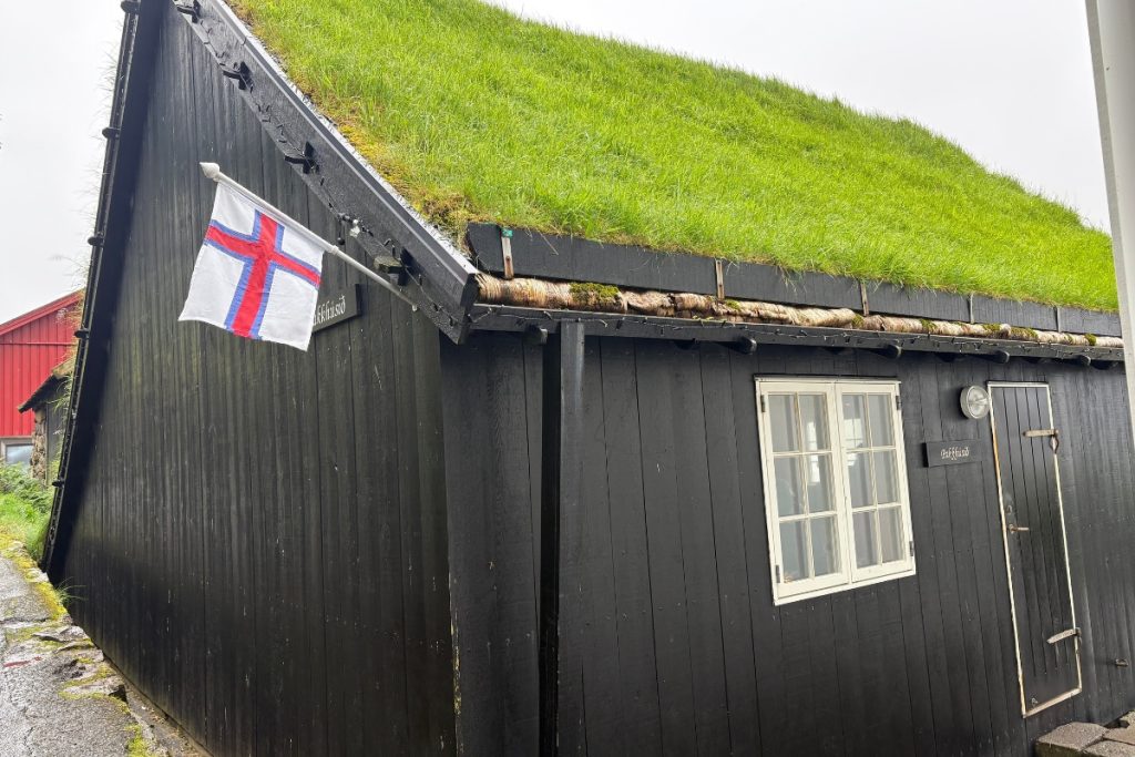 A black timber building with a grass roof and Faroese flag, the Pakkhúsið café in Bøur