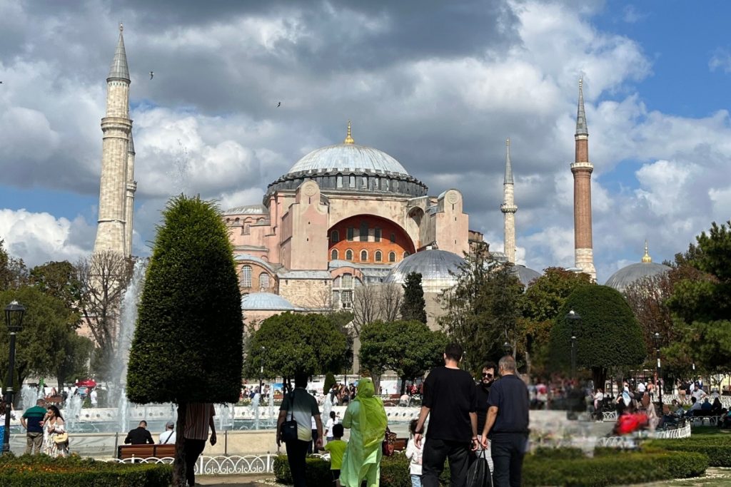 Wide view of Hagia Sophia's exterior with multiple domes and four minarets, seen across a park with a fountain, trees, and visitors in the foreground.