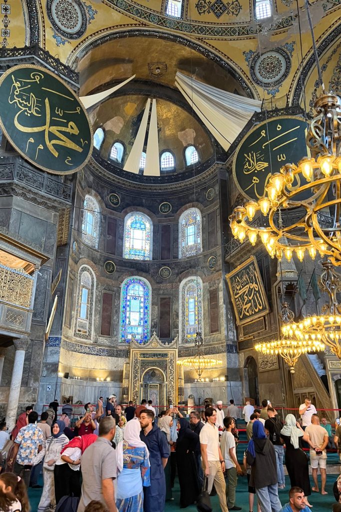 Interior view of Hagia Sophia's apse showing the gold mihrab niche flanked by tall stained glass windows, with crowds of visitors gathered below.