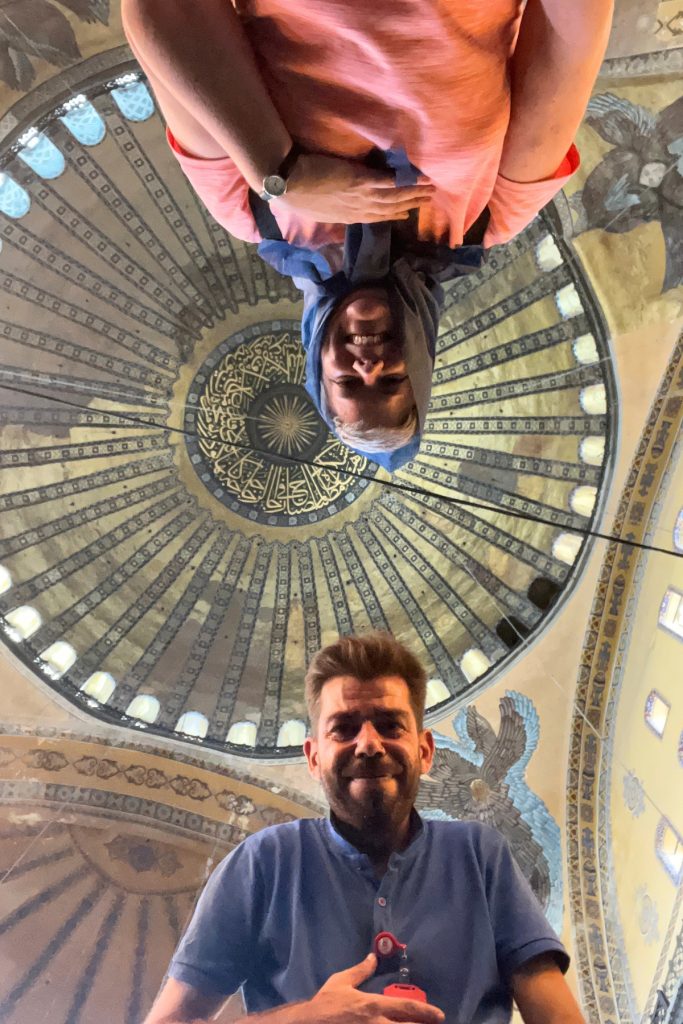 Upside-down photo looking up at Hagia Sophia's dome interior, with two people's faces visible at the top and bottom of the frame during a selfie attempt.