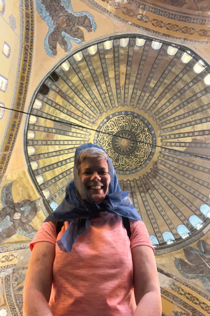 Woman smiling while photographing Hagia Sophia's dome ceiling from below, with intricate gold and blue medallion artwork visible overhead.