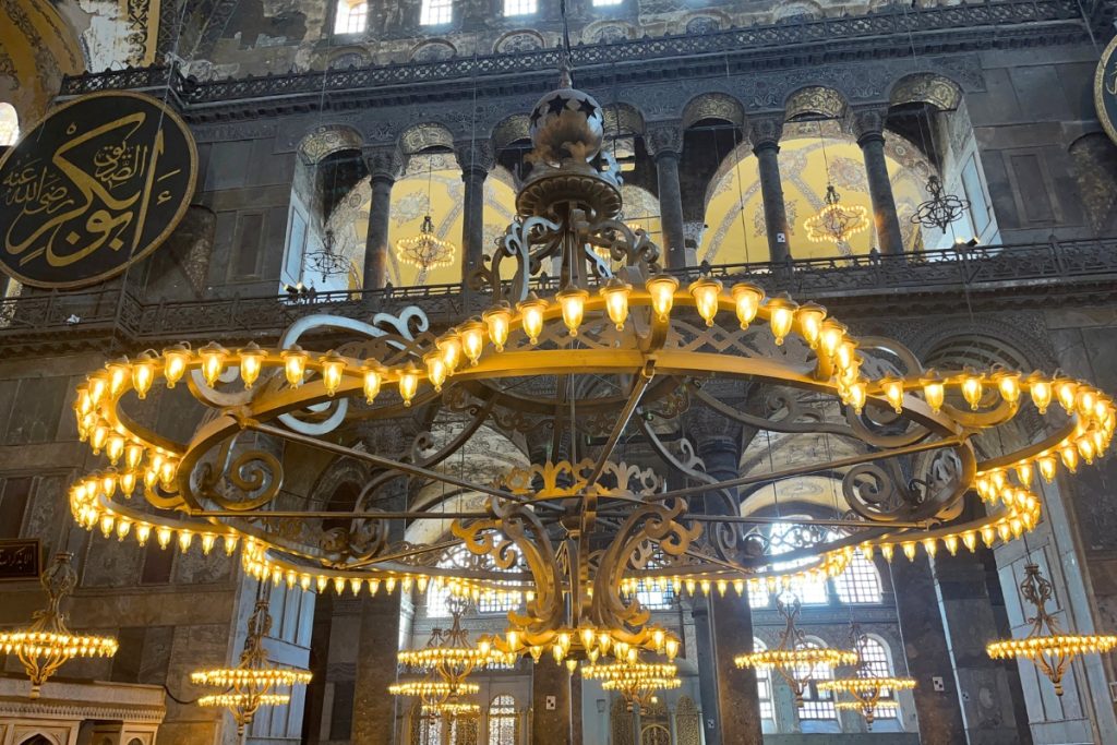 Large gold chandelier hanging inside Hagia Sophia beneath an ornately decorated dome with Arabic calligraphy medallions and archways in the background.