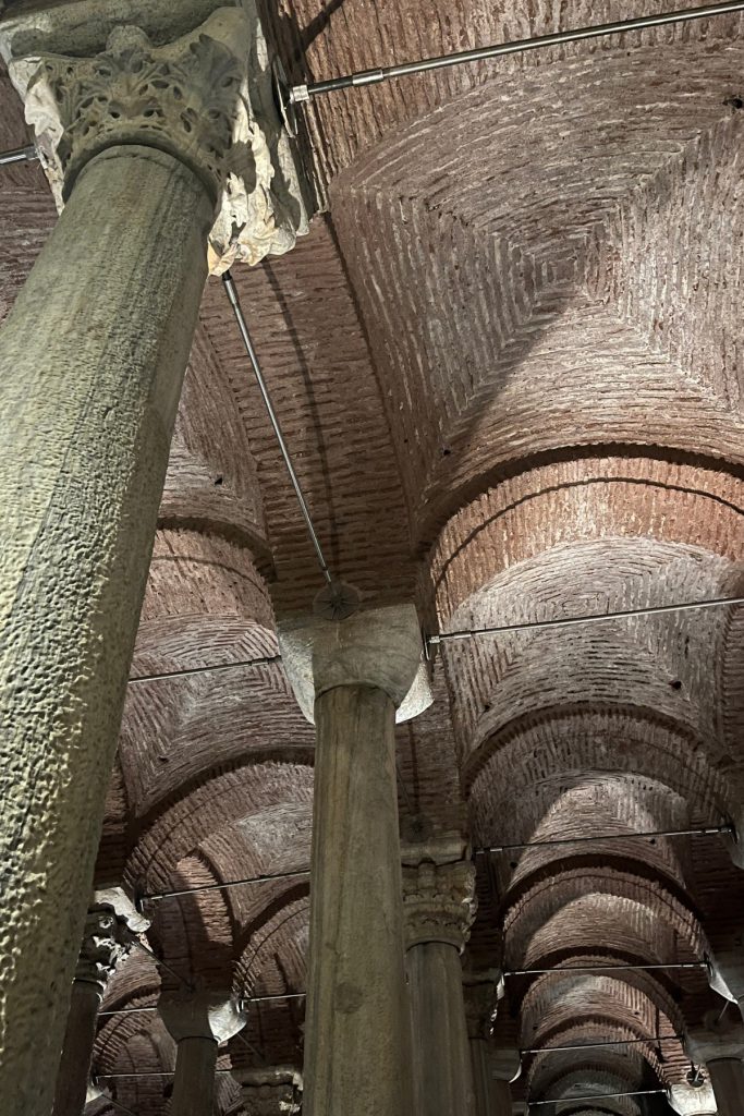 Close-up looking up at a stone column capital and intersecting brick vaulted ceiling inside the Basilica Cistern.