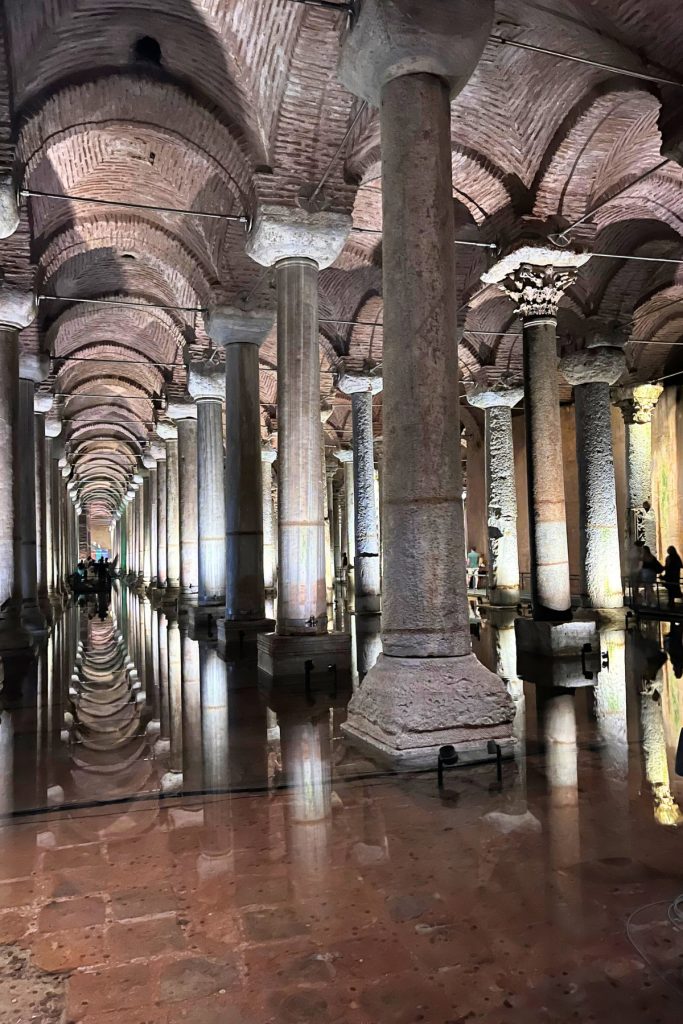 Rows of weathered stone columns with brick vaulted ceilings above, reflected in shallow water inside the Basilica Cistern, with visitors visible in the background.