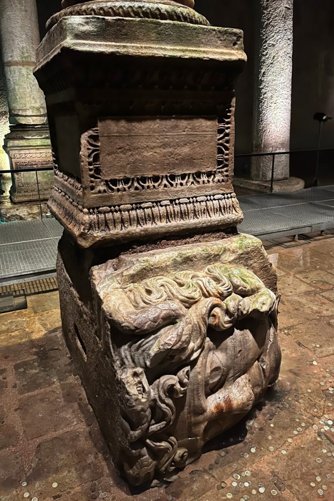 Carved stone Medusa head positioned sideways as the base of a column inside the Basilica Cistern, with coins scattered on the ground nearby.