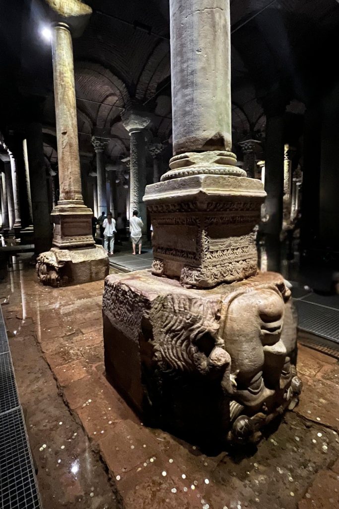 Carved stone Medusa head positioned upside down as the base of a column inside the Basilica Cistern, with a visitor standing in the background.