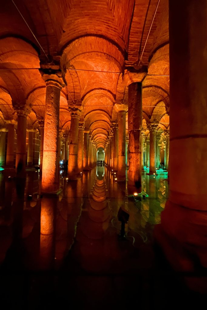 Rows of stone columns reflected in still water, lit in warm amber and orange, with a long corridor stretching into the distance inside the Basilica Cistern.