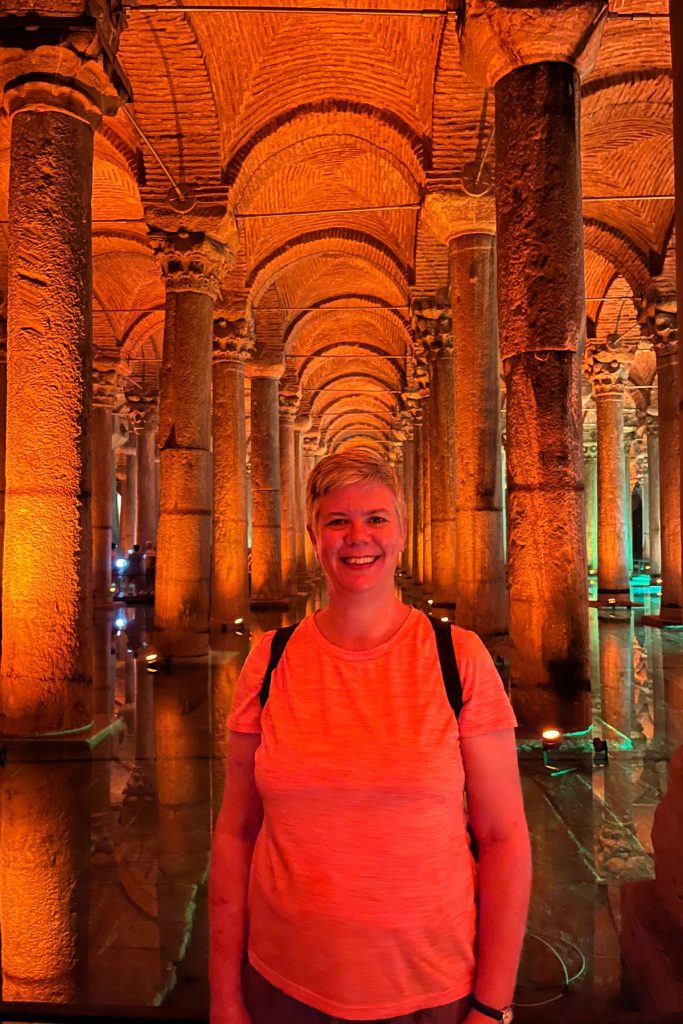 Woman smiling in an amber-lit corridor of stone columns reflected in water inside the Basilica Cistern.