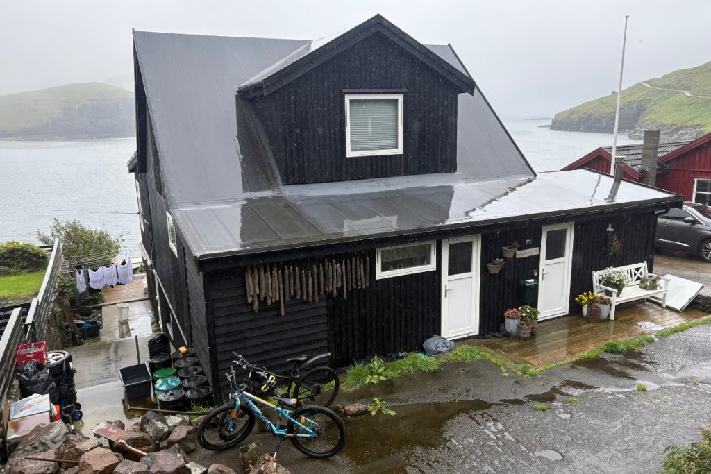 A black timber house with bicycles and laundry drying outside, overlooking the fjord in Bøur