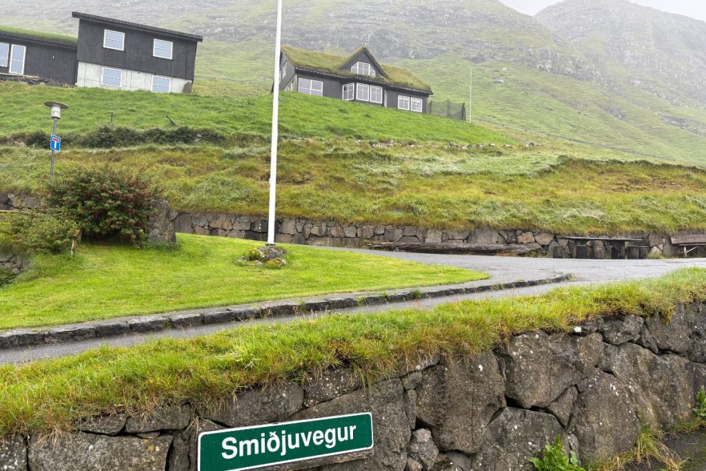 A green street sign reading Smiðjuvegur with black turf-roofed houses on a hillside in Bøur, Faroe Islands