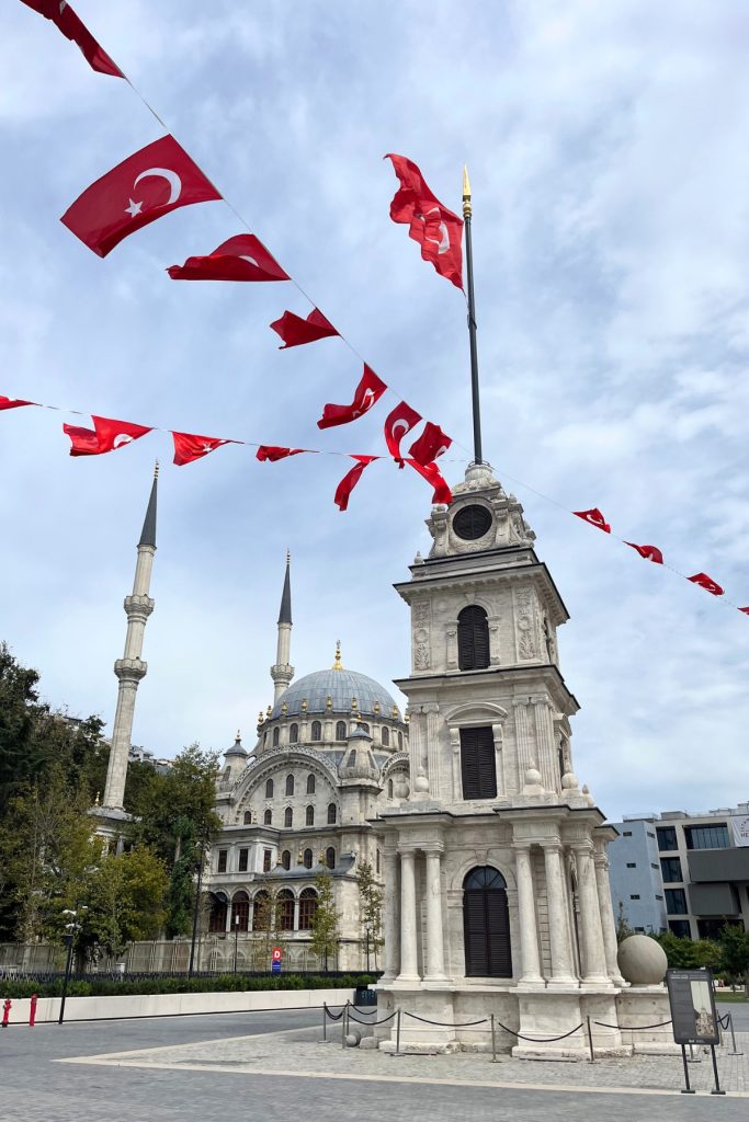 Ornate white mosque with twin minarets and a domed clock tower, framed by rows of Turkish flags on a cloudy day.
