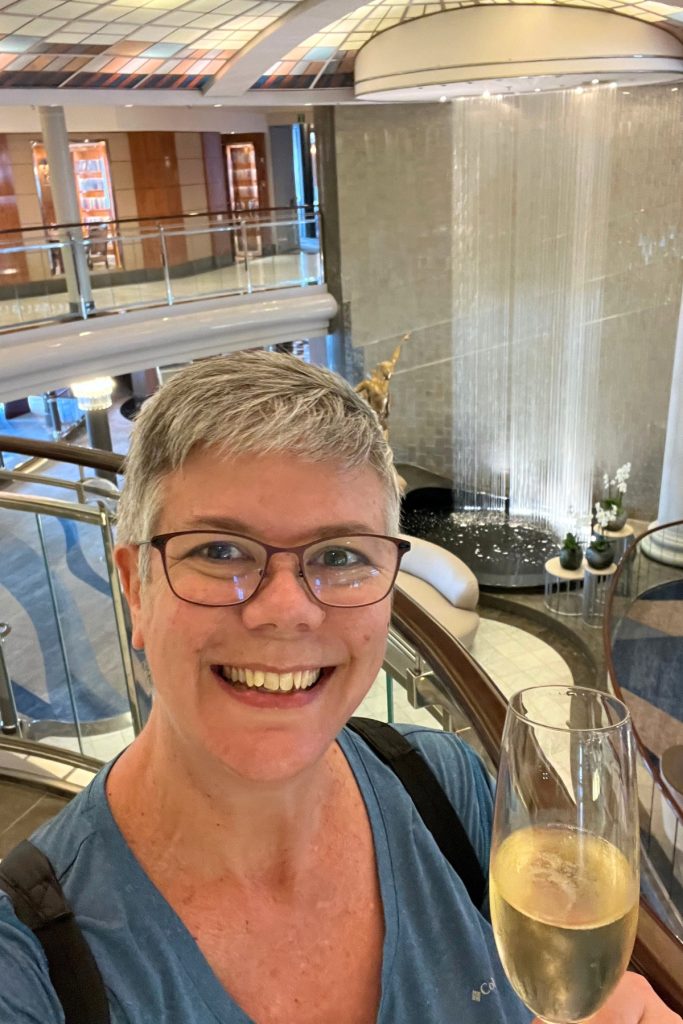 Selfie on an upper deck balcony inside the ship's multi-story atrium, holding a glass of champagne, with a rainfall-style water feature visible below.