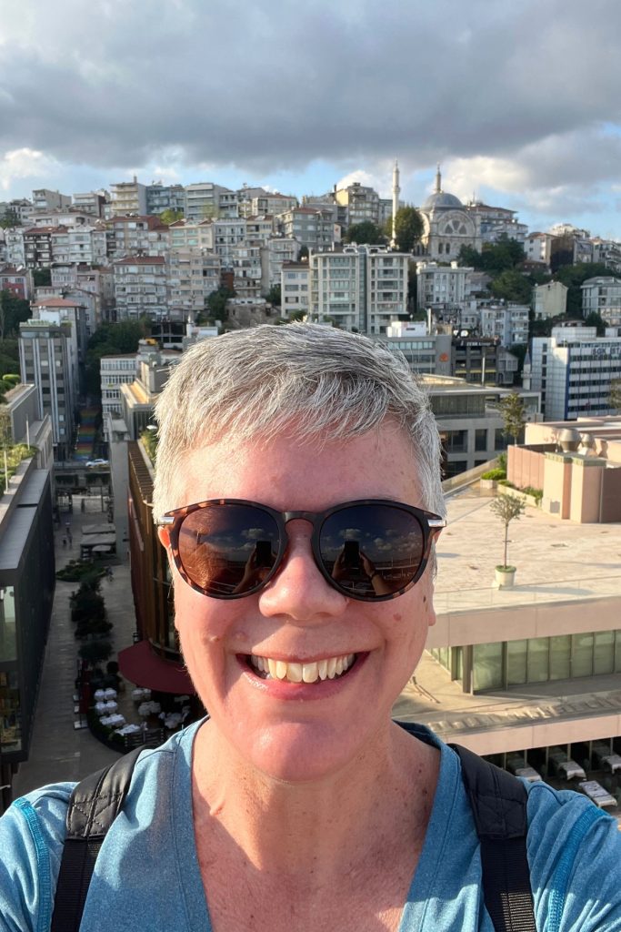 Selfie with a dense hillside neighborhood of apartment buildings and a domed mosque with two minarets visible in the background under a partly cloudy sky.