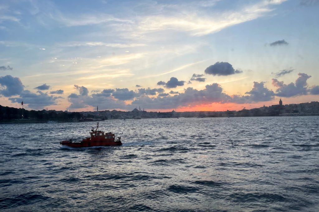 Small red boat crossing the water at sunset with the Istanbul skyline silhouetted in the distance under a colorful sky.