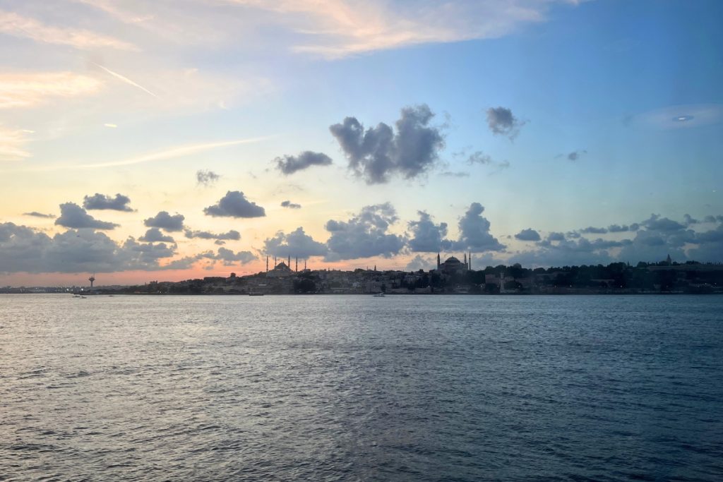 Silhouette of the Istanbul skyline at sunset, including mosque domes and minarets, seen across the water under an orange and blue sky.