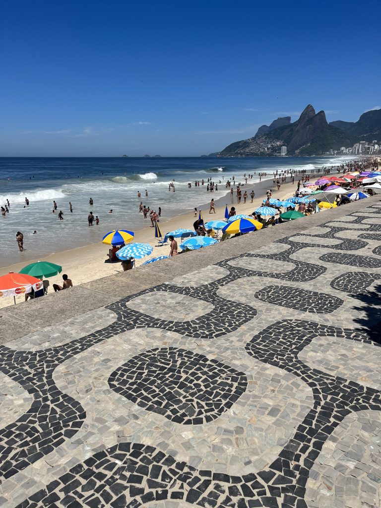 Wide view of Ipanema Beach with the calçada portuguesa mosaic sidewalk, umbrellas, and the Dois Irmãos peaks in Rio de Janeiro
