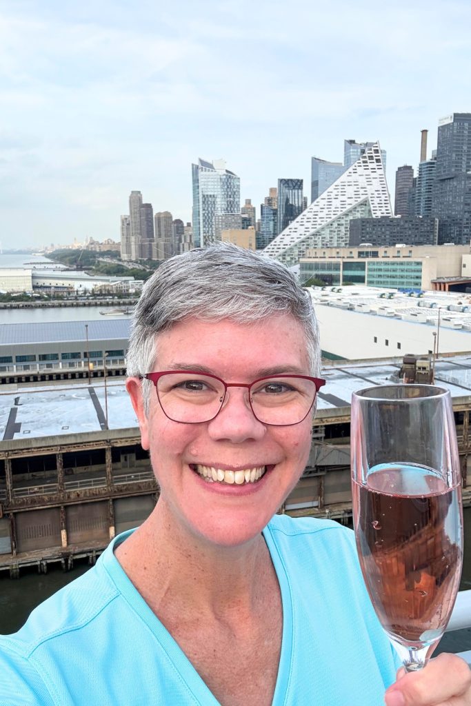 Smiling woman holding a glass of champagne with the Manhattan skyline in the background during sail-away aboard Virgin Voyages Brilliant Lady