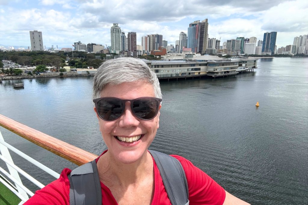 Woman on the upper deck of a cruise ship departing Manila, with the Rizal Park flagpole visible across the water among the city skyline