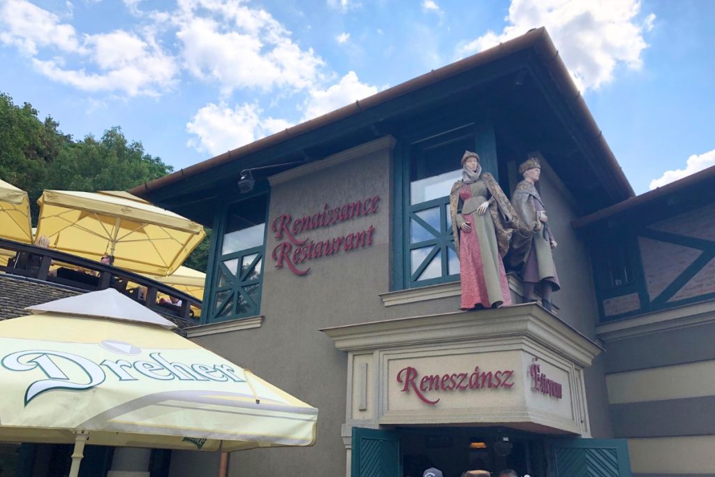 Exterior of the Renaissance Restaurant in Hungary, with costumed mannequin figures on the upper balcony and yellow umbrellas on the terrace below.