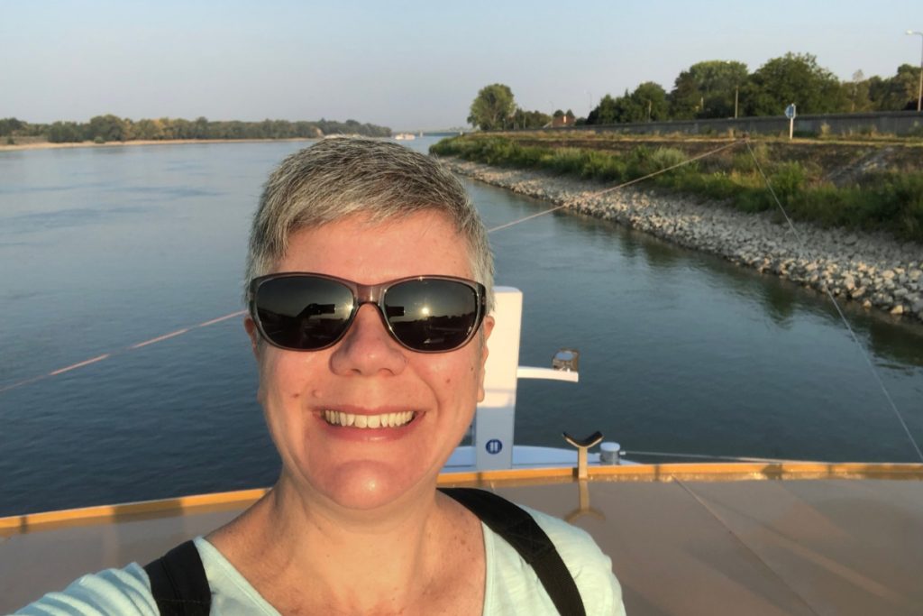 Selfie on the deck of a river cruise ship docked near Komárno, Slovakia, with the Danube River and a low, rocky bank behind.