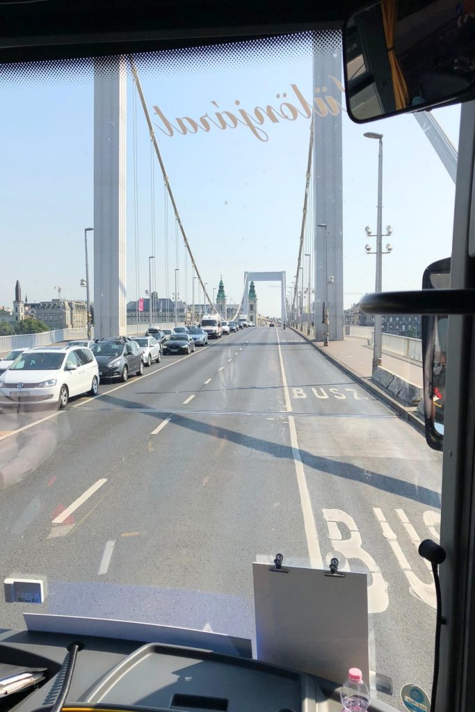 View from inside a tour bus crossing a suspension bridge into Budapest, with cars queued ahead.