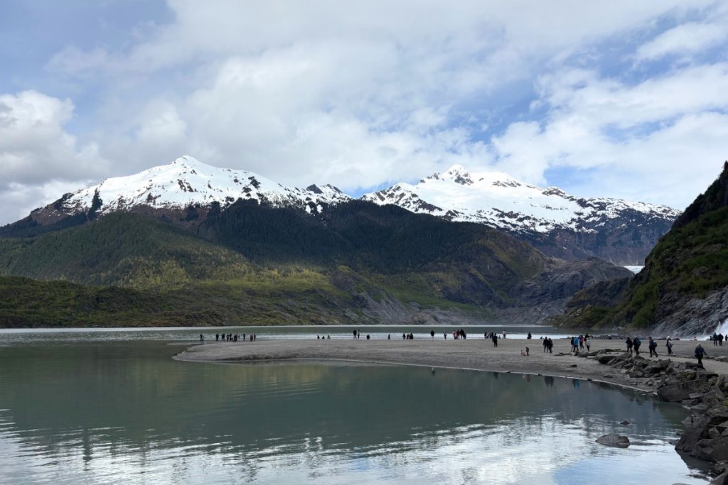 A wide view of Mendenhall Glacier between snow-capped mountains, with visitors gathered on a gravel beach along the lake in the foreground.