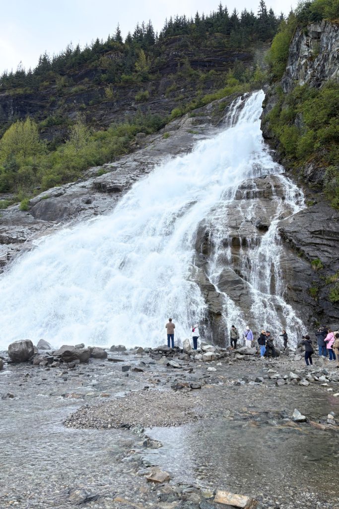 Hikers standing on a rocky beach photographing Nugget Falls as it drops into Mendenhall Lake.