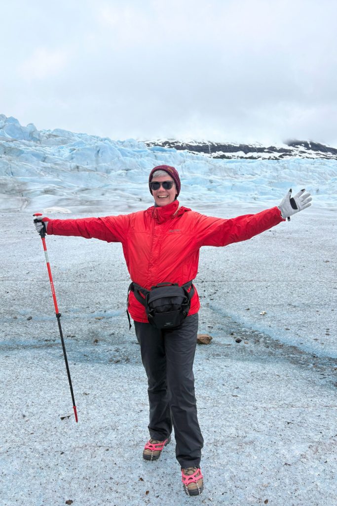 A woman in a red jacket, sunglasses, and crampons standing with arms outstretched on Mendenhall Glacier, holding a hiking pole.