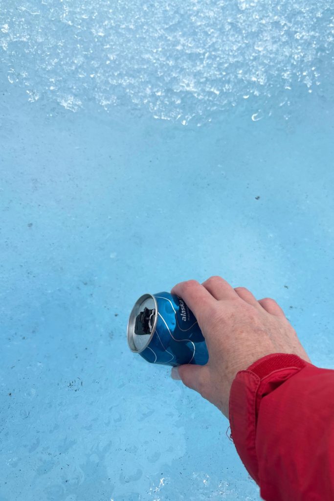 A hand in a red sleeve holding an empty blue water can against the icy blue surface of Mendenhall Glacier.