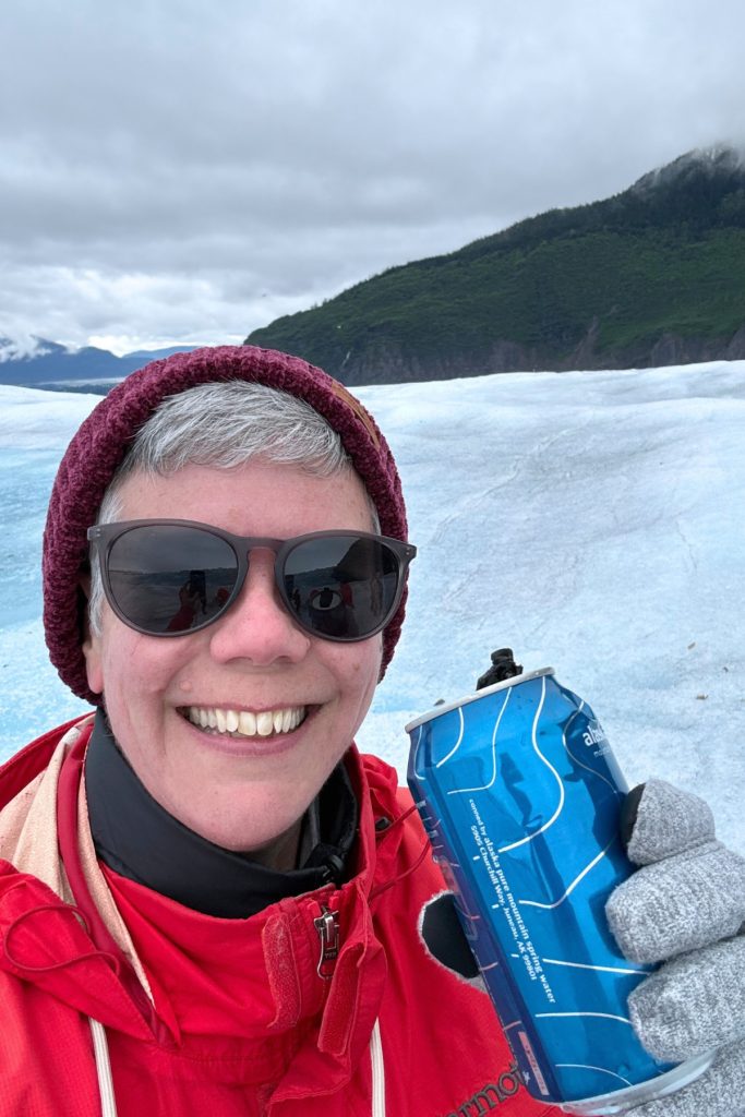 A woman in sunglasses and a red jacket smiling while holding a blue water can on the surface of a glacier.