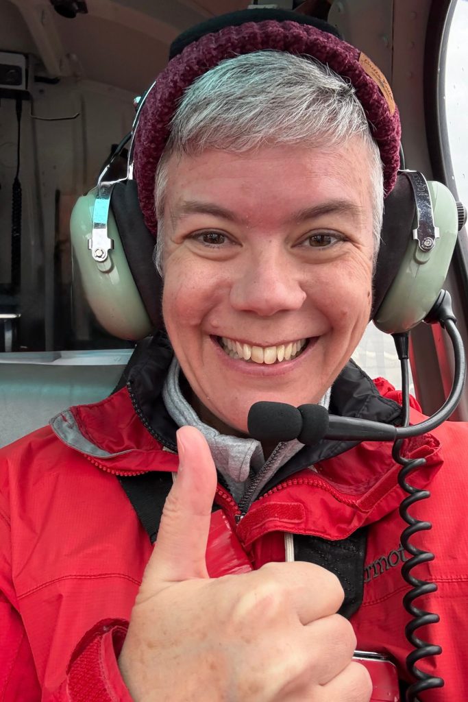 A woman in a red jacket wearing a helicopter headset and giving a thumbs up inside the cabin before takeoff.