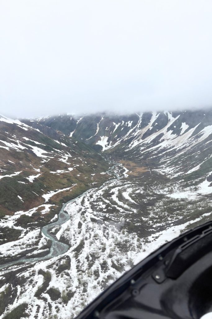 Aerial view from a helicopter of a winding glacial river cutting through a snow-patched mountain valley in Juneau, Alaska.