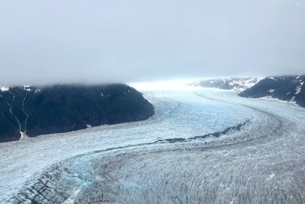 Aerial view of Mendenhall Glacier's crevassed ice field winding between dark mountain ridges under a cloudy sky.