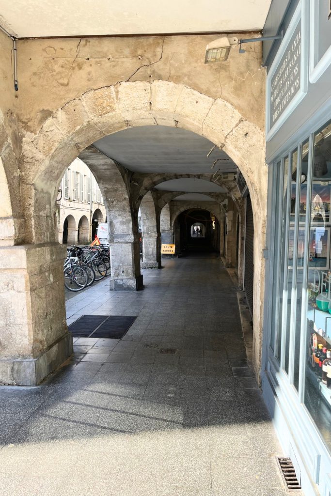 Stone arcade passageway in La Rochelle, France, with parked bicycles, a storefront, and a figure visible at the far end of the covered walkway.