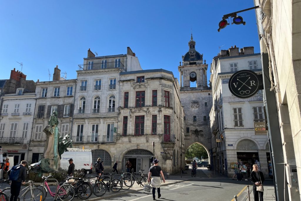 Street view in La Rochelle, France, looking toward the Tour de la Grosse Horloge clocktower gate, with a bronze statue, parked bicycles, and pedestrians in the foreground.