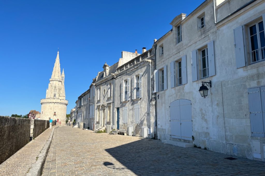 Cobblestone rampart walkway in La Rochelle, France, lined with pale stone townhouses, leading toward the spired Tour de la Lanterne in the distance.