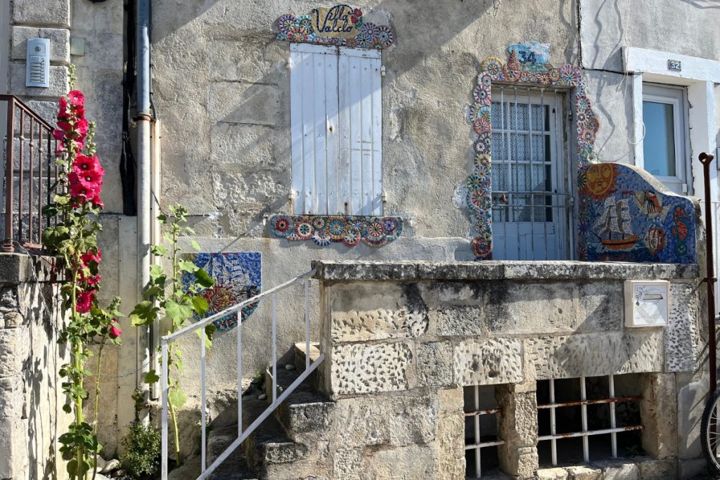Stone house facade in La Rochelle, France, decorated with colorful mosaic tile work around the windows, with red hollyhocks growing up the wall.
