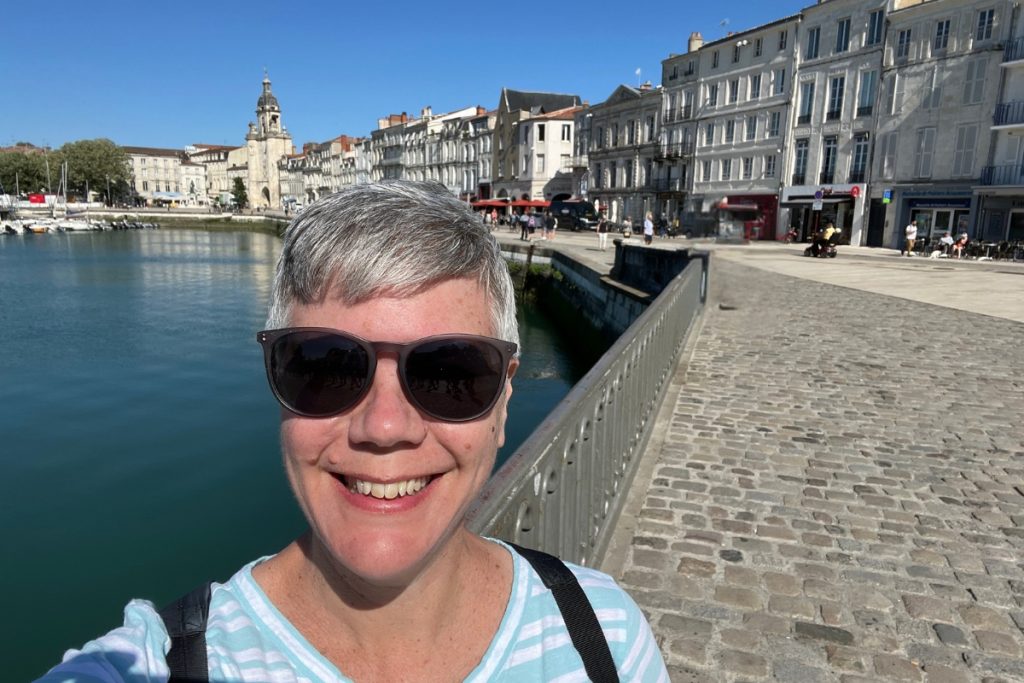 Bethany smiling in sunglasses on a bridge over the Vieux Port in La Rochelle, France, with the harbor, Grosse Horloge clocktower, and waterfront buildings behind her.