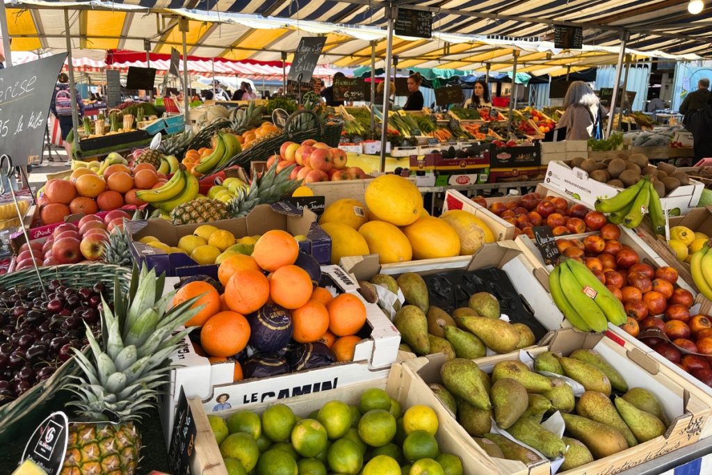 Outdoor farmers market stalls in La Rochelle, France, piled with fresh fruit including pineapples, oranges, melons, and pears under striped awnings.