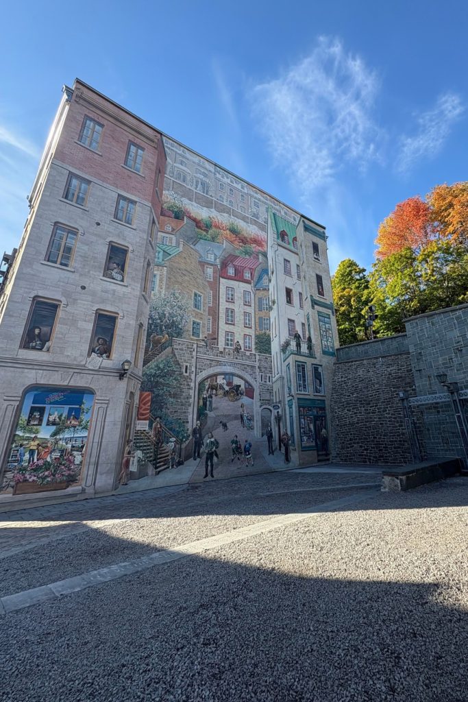 Trompe-l'oeil mural in Quebec City depicting layered building facades, painted figures in windows, and a staircase, with fall foliage beside the wall.