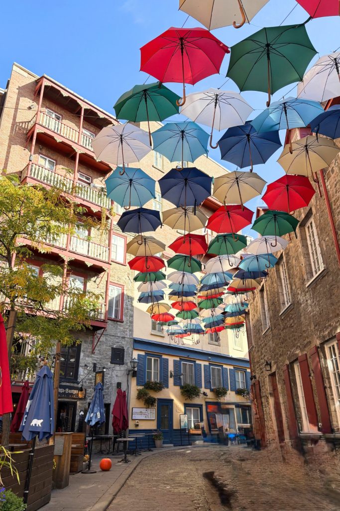 Colorful umbrellas suspended above a cobblestone street in Quebec City's Petit-Champlain district, with cafe patios below.