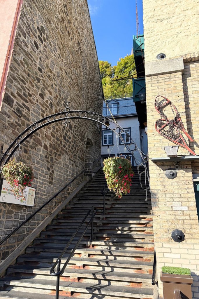 Stone staircase in Quebec City with a wrought-iron arch, hanging flower baskets, and a snowshoe mounted on the brick wall beside it.