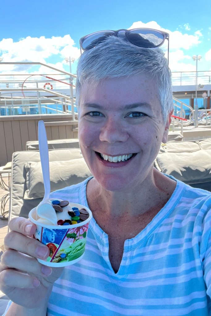 Bethany smiling on the pool deck of a cruise ship, holding a cup of soft serve ice cream topped with M&Ms, sunglasses pushed up on her head.
