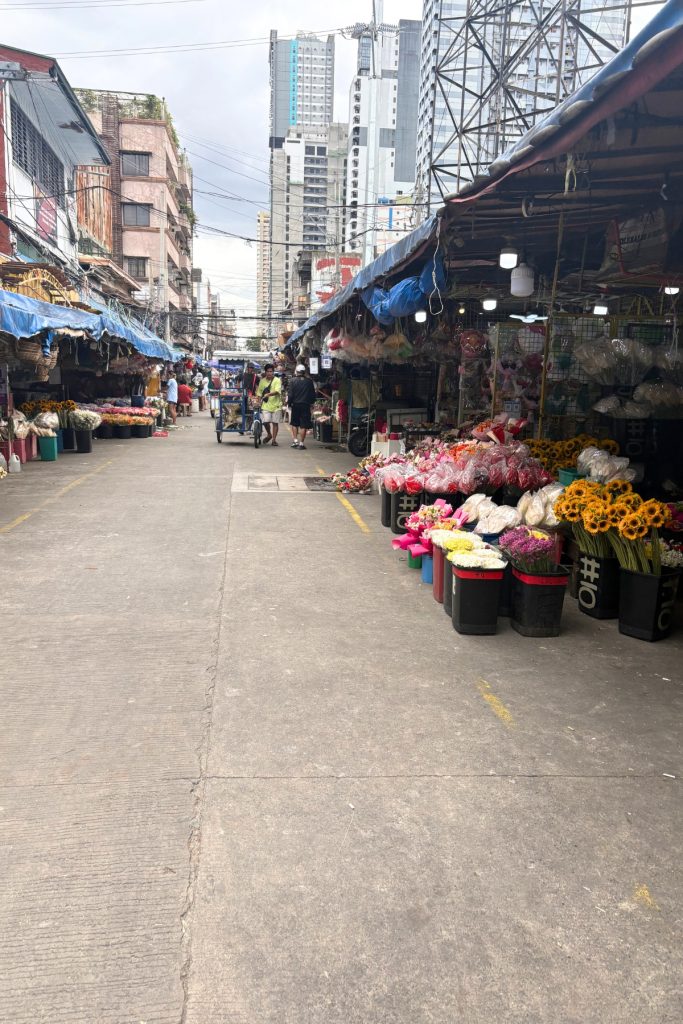 Street lined with flower stalls and buckets of sunflowers and carnations at Dangwa Market in Manila, with the city skyline behind