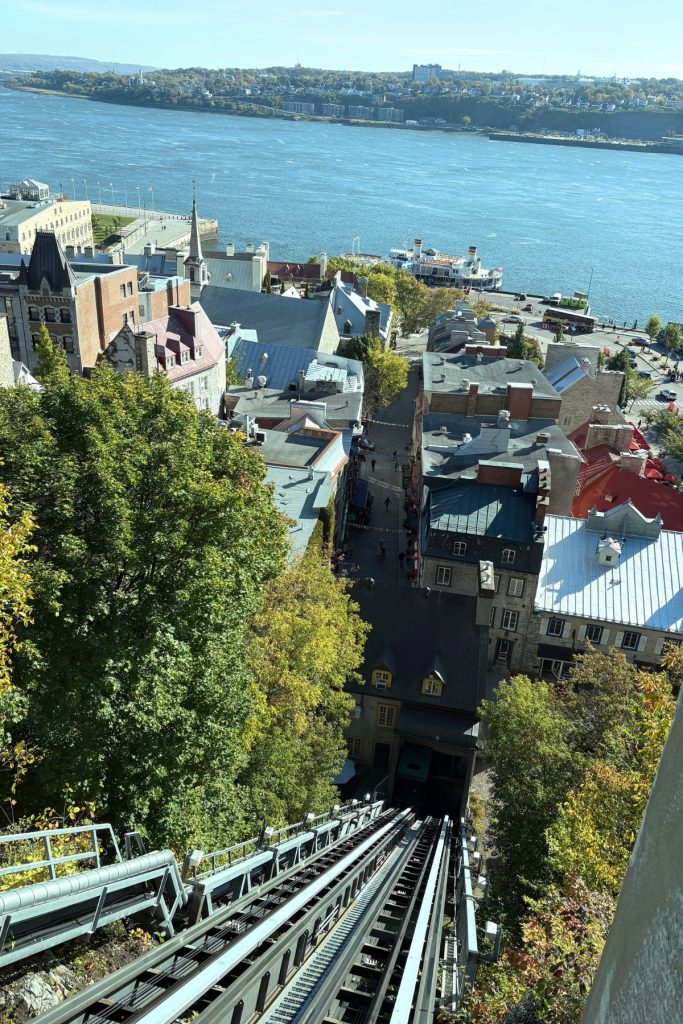 View straight down the funicular tracks in Quebec City, overlooking rooftops, a cobblestone street, and the St. Lawrence River with a docked riverboat.