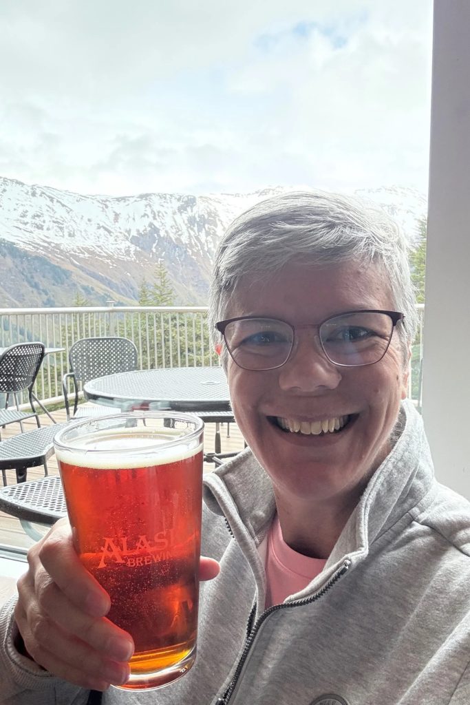 A woman holding a pint of amber beer with snow-capped mountains visible through a restaurant window at the Mount Roberts tram summit.