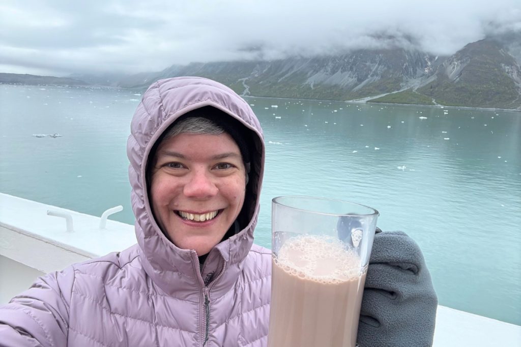 Smiling woman in a winter coat holding a warm drink aboard a ship in Alaska, with icebergs and mountains in the background