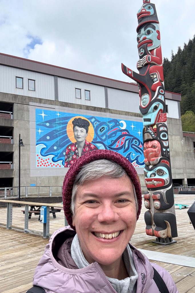 A woman smiling in front of a three-story mural of Elizabeth Peratrovich and a totem pole at Sealaska Plaza in Juneau
