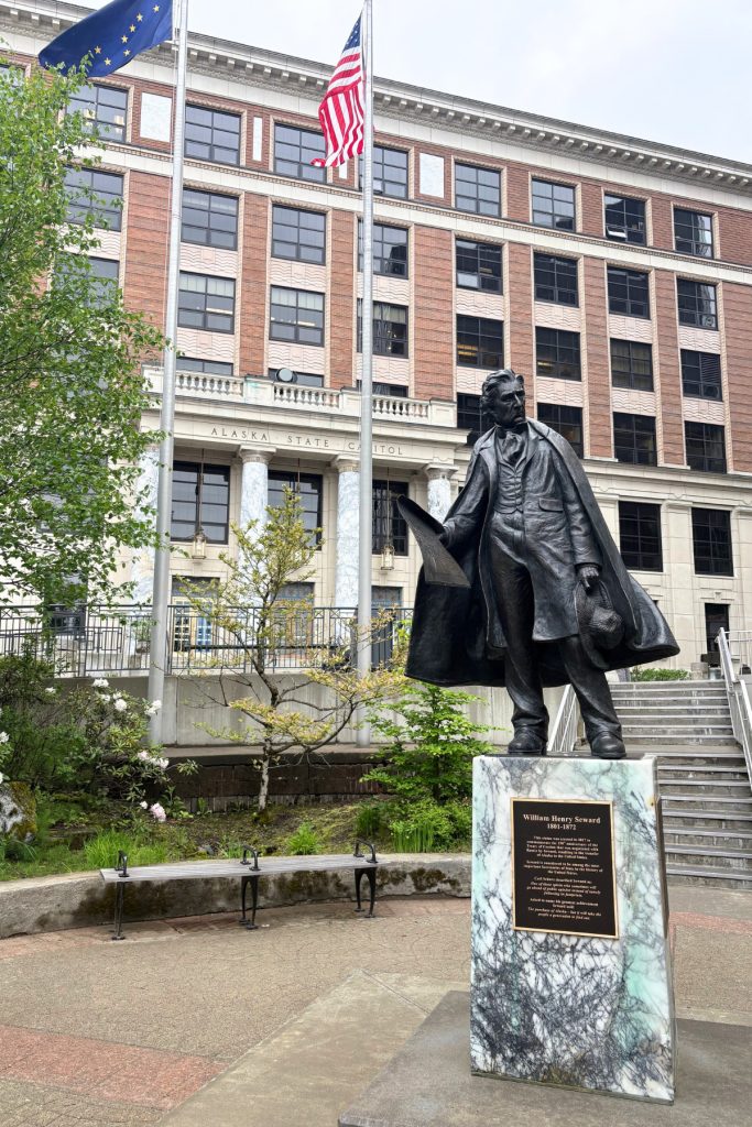 A bronze statue of William Henry Seward holding his hat, standing on a marble pedestal in front of the Alaska State Capitol.